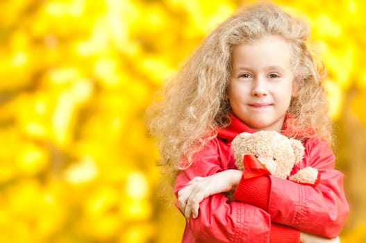Beautiful and happy little girl with teddy bear on her hands. Smiling and looking into the camera. Yellow autumn trees in background.