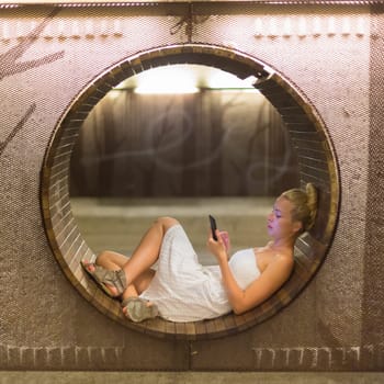 Lady wearing white summer dress playing carelessly with her android smart phone browsing trough social networks relaxing on contemporary circular bench in urban pedestrian underpass.