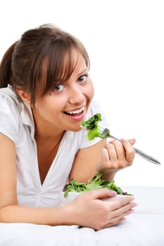 Young woman eating healthy salad isolated on white background lying on the bed