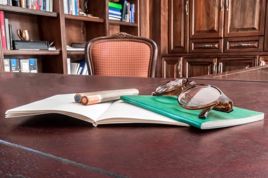 A private study in a residential home with an up close view of a set of two notebooks, two pencils and a pair of reading glasses on the desk.