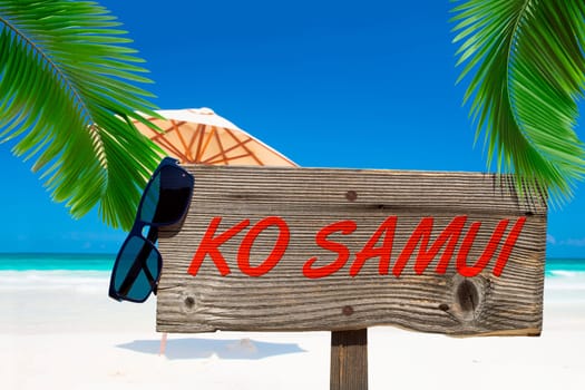 Wooden sign with the inscription "Ko Samui"" under palm trees on the sandy beach"