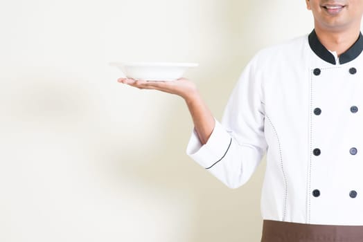 Portrait of Indian male chef in uniform presenting an empty dish, standing on plain background with shadow, copy space.