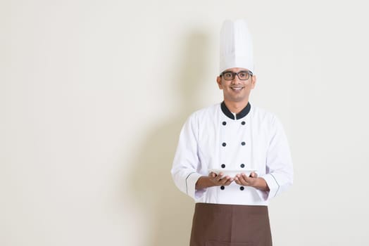 Portrait of handsome Indian male chef in uniform hands holding an empty plate and smiling, standing on plain background with shadow, copy space.