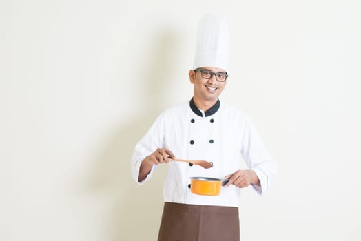 Portrait of handsome Indian male chef in uniform holding cooking pot and stirring food, standing on plain background with shadow, copy space on side.