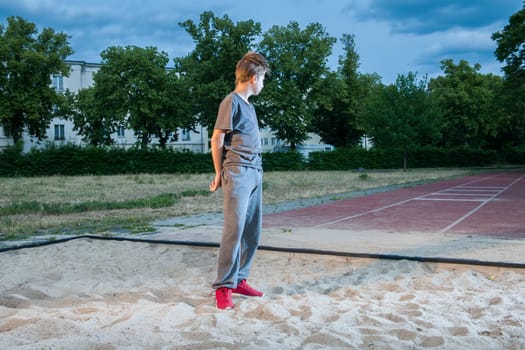 Side full-body view of a young male teenager standing in a diving pit looking at the start-up track with arms placed on his back