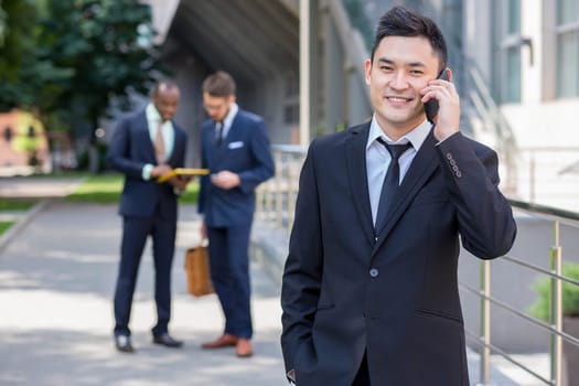 Portrait of multi ethnic business team. Three happy smiling men standing against the backdrop of the city.  The foreground of a Chinese  man  with phone