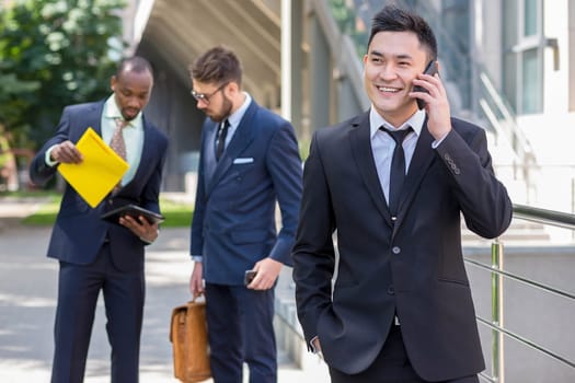 Portrait of multi ethnic business team. Three happy smiling men standing against the backdrop of the city.  The foreground of a Chinese  man  with phone