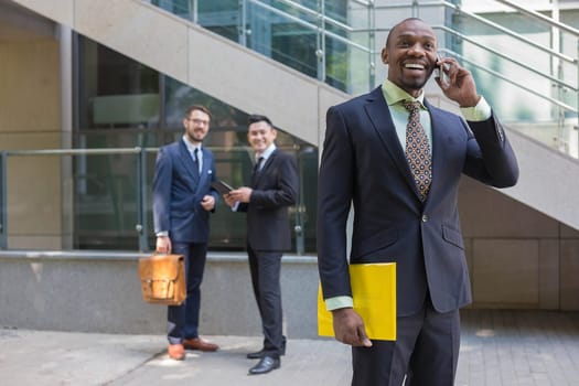 Portrait of multi ethnic business team. Three happy smiling men standing against the backdrop of the city.  The foreground of a African-American man talking on the phone
