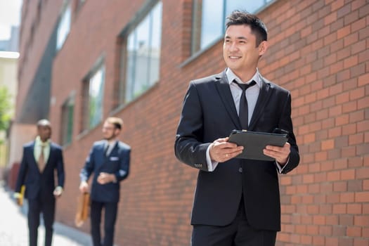 Portrait of multi ethnic business team. Three happy smiling men standing against the backdrop of the city.  The foreground of a Chinese  man  with phone and laptop