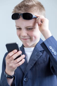 Young boy dressed up as a spy. He is looking at a smartphkone and pulling a funny face with his glasses being held off his eyes. He is wearing a suit and is in front of a plain background