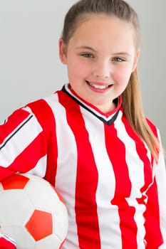 Young girl posing for the camera in a football kit with a football under her arm. She is smiling for the camera against a plain background.