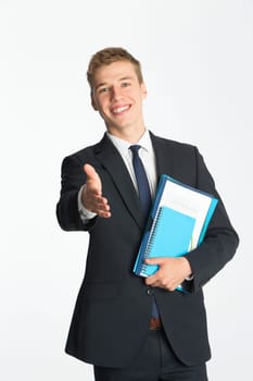 Young businessman holding his hand out to offer a handshake. He is dressed in a suit and is holding folders whilst standing against a plain background