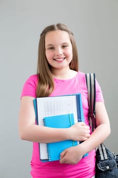 Young girl posing for the camera with her school folders and her bag. She is standing against a plain background.