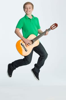 Young boy jumping whilst playing guitar. He is smiling at the camera and is against a plain background.