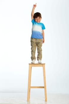 Little boy standing on a high stool, pointing up to the ceiling. He is against a white background.