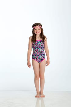 Full length shot of a little girl wearing a swimming costume with goggles on her head. She is smiling at the camera and standing against a white background.