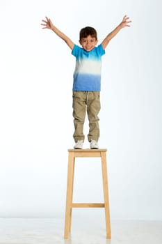 Little boy standing on a high stool with his arms out stretched. He is against a white background.