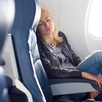 Tired blonde casual caucasian lady napping on uncomfortable seat while traveling by airplane. Commercial transportation by planes.