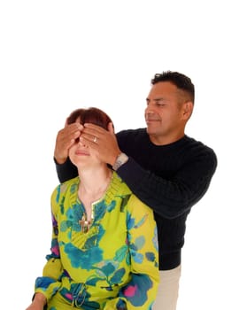 A Hispanic man holding his hands over the eye's of his Caucasian wife,.standing behind her, isolated for white background..