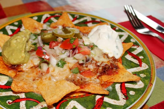 A plate of delicious tortilla nachos with melted cheese sauce, ground beef, jalapeno peppers, onions, tomato, cheese, salsa, and sour cream with guacamole dip
