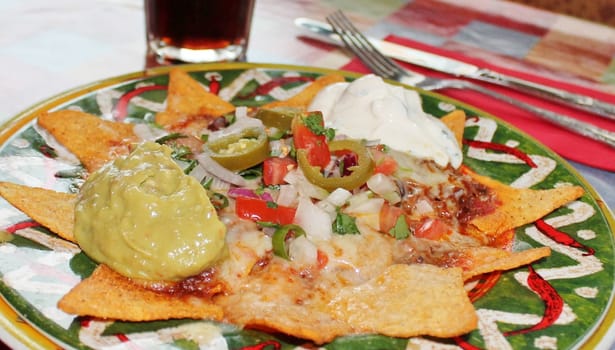 A plate of delicious tortilla nachos with melted cheese sauce, ground beef, jalapeno peppers, onions, tomato, cheese, salsa, and sour cream with guacamole dip