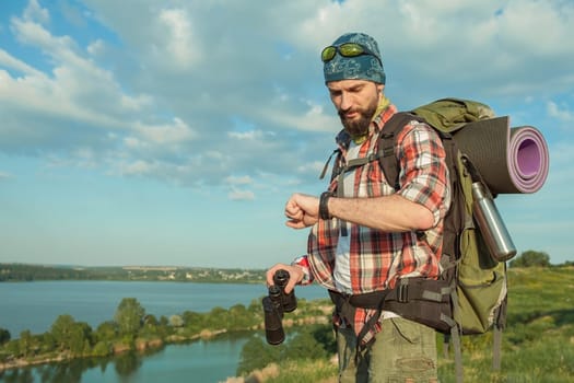 Young caucasian tourist with backpack  standing on the top of hill and looking at  watch. The concept of an invitation to travel