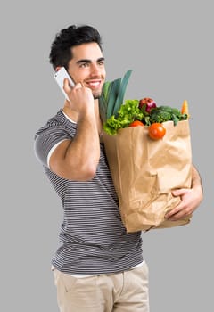 Handsome young man carrying a bag full of vegetables and making a selfie, isolated over gray background