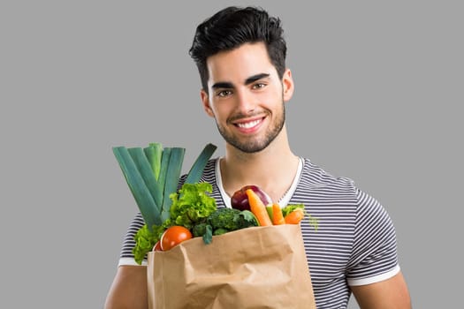 Handsome young man carrying a bag full of vegetables, isolated over gray background