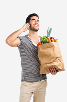 Handsome young man carrying a bag full of vegetables and making a phone call, isolated over gray background