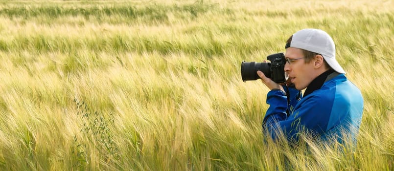 Wide format image of a photographer sitting amidst beautiful barley plants on a field and aiming the camera