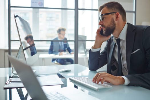 Modern businessman consulting partner by phone while sitting in front of computer in office