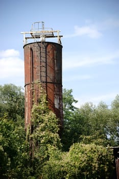 An old rusted storage tank or silo is overgrown by bushes.