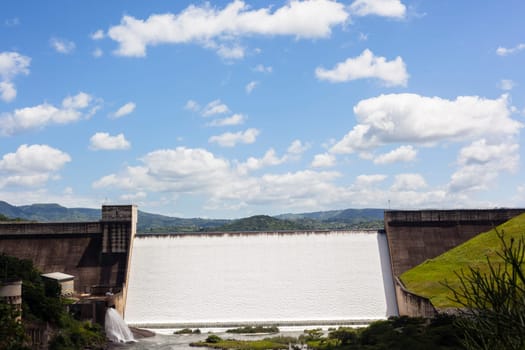 Dam wall full flowing river water over weir wall into summer valley.