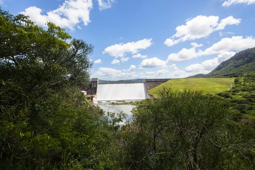 Dam wall full flowing river water over weir wall into summer valley.