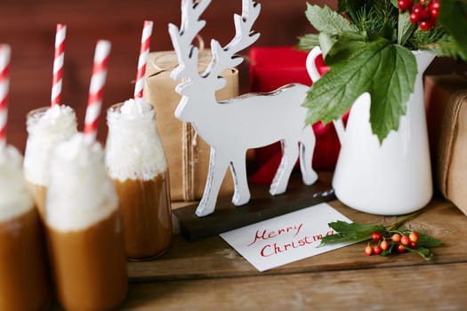 Wooden deer, porcelain vase with guelder leaves and berries, bottles with hot chocolate with whipped cream and gifts on table