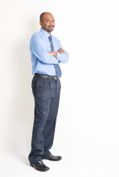 Portrait of full body mature Indian business man with blue shirt arms crossed standing on plain background.