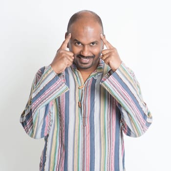 Portrait of mature casual Indian man thinking, fingers pointing on head, standing on plain background.