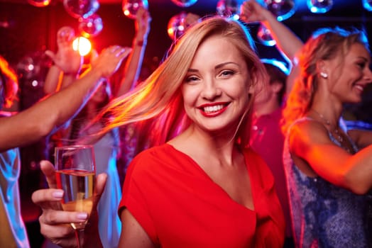 Joyful girl with flute of champagne looking at camera in night club with her friends dancing on background