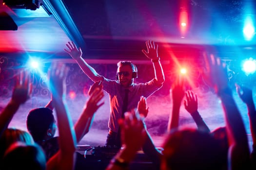 Energetic deejay with headphones having fun by turntables in front of dancing crowd