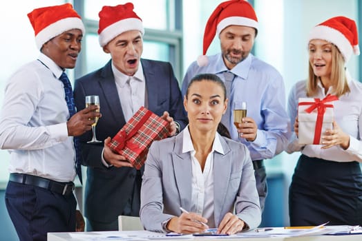 Amazed businesswoman looking at camera at workplace while colleagues with gifts and champagne congratulating her behind