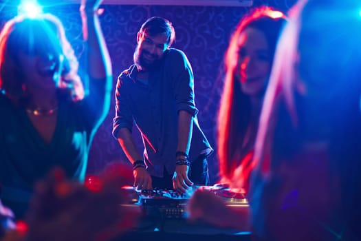 Happy bearded deejay standing by turntables in front of dancing crowd and looking at camera