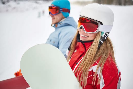 Happy young snowboarder in activewear looking at camera on background of her boyfriend on winter resort