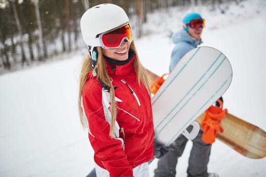 Young female with snowboard looking at camera on background of her boyfriend on winter resort