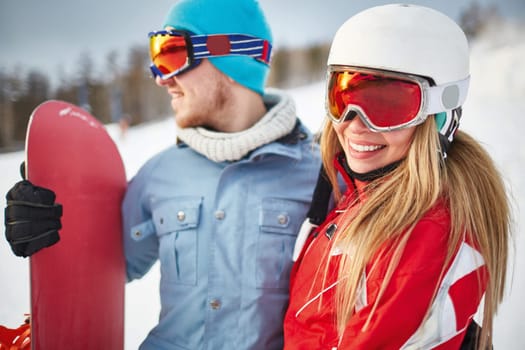 Pretty female snowboarder in winter activewear looking at camera during vacation at sportive resort with her boyfriend near by