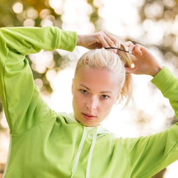 Portrait of attractive confident sporty woman wearing fashionable green sportswear  making her hair in ponytail before training in nature. Active sporty lifestyle concept.