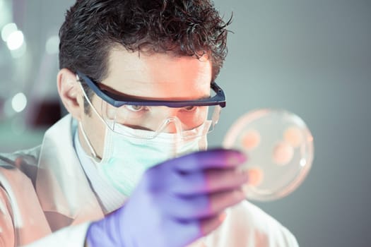 Lab coat, mask and goggles protected life science researcher observing potentially infectious cells in petri dish. Focus on scientist&#39 s eye. Healthcare and biotechnology concept.