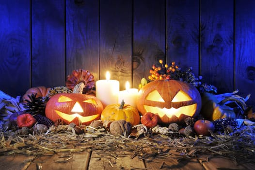 illuminated Halloween pumpkins,candles,nuts,corn cobs and apple on straw in front of old weathered wooden boards in the blue evening light
