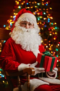 Happy Santa with box looking at camera while sitting by sparkling firtree