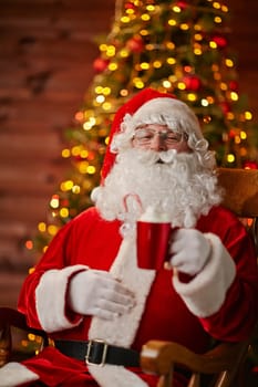 Joyful Santa with cup of latte sitting in armchair in front of camera