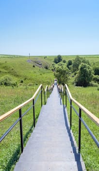 Metal staircase with handrails leading down to the holy spring. Large Orthodox cross on a hill.